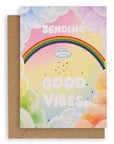Colorful greeting card with cumulus clouds surrounding a rainbow with a UFO below it, followed by rainbow stars and clouds, and the words "Sending Good Vibes" on the front. Shown with a brown kraft paper envelope.