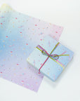 Single sheet of gift wrap with pastel gradient background and neon icons. Shown wrapped on present with pink and green velvet ribbon.