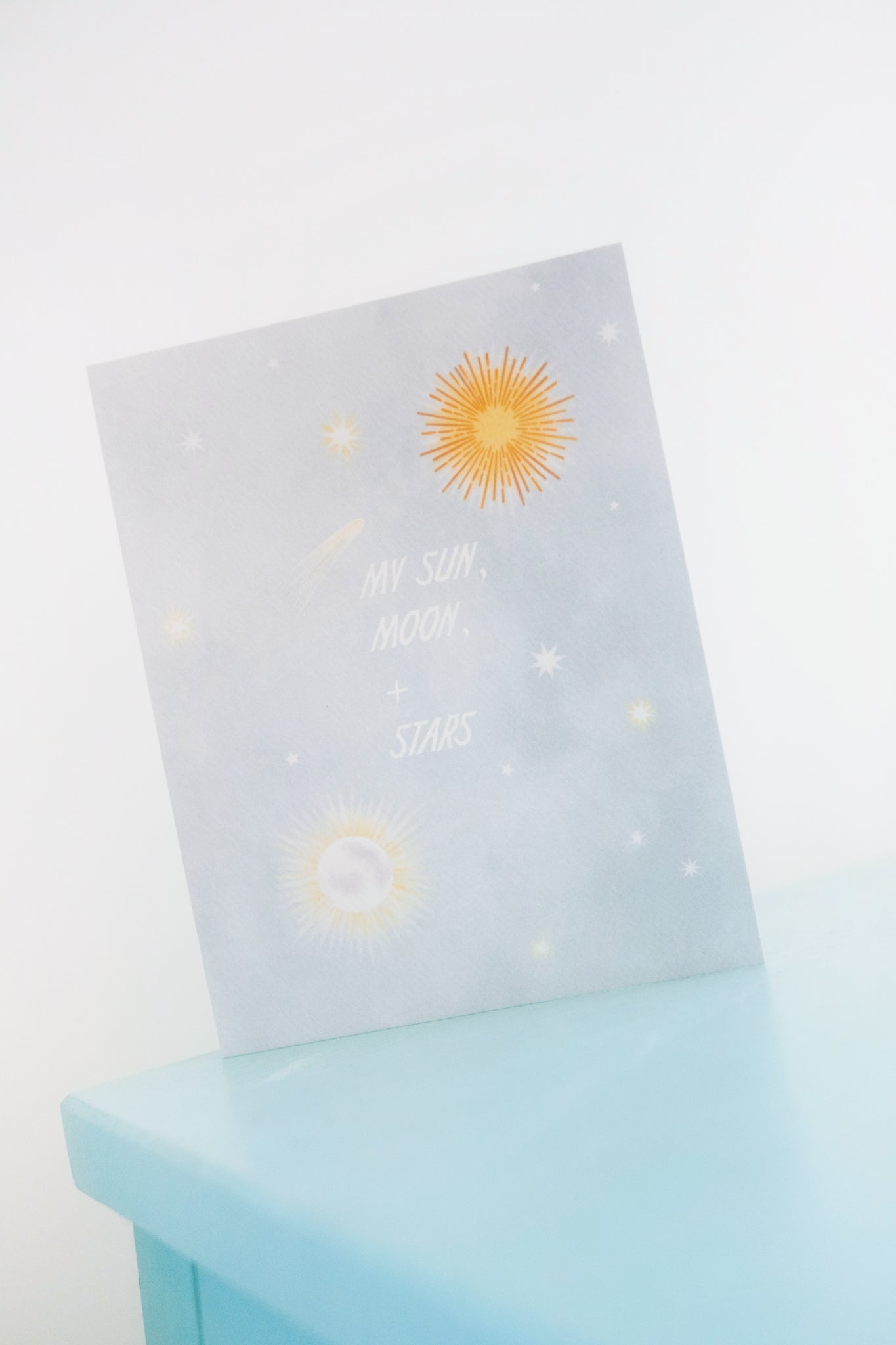 Blue sky with sun, moon, stars and text that reads &quot;My Sun, Moon, + Stars.&quot; Shown on blue shelf with white background.