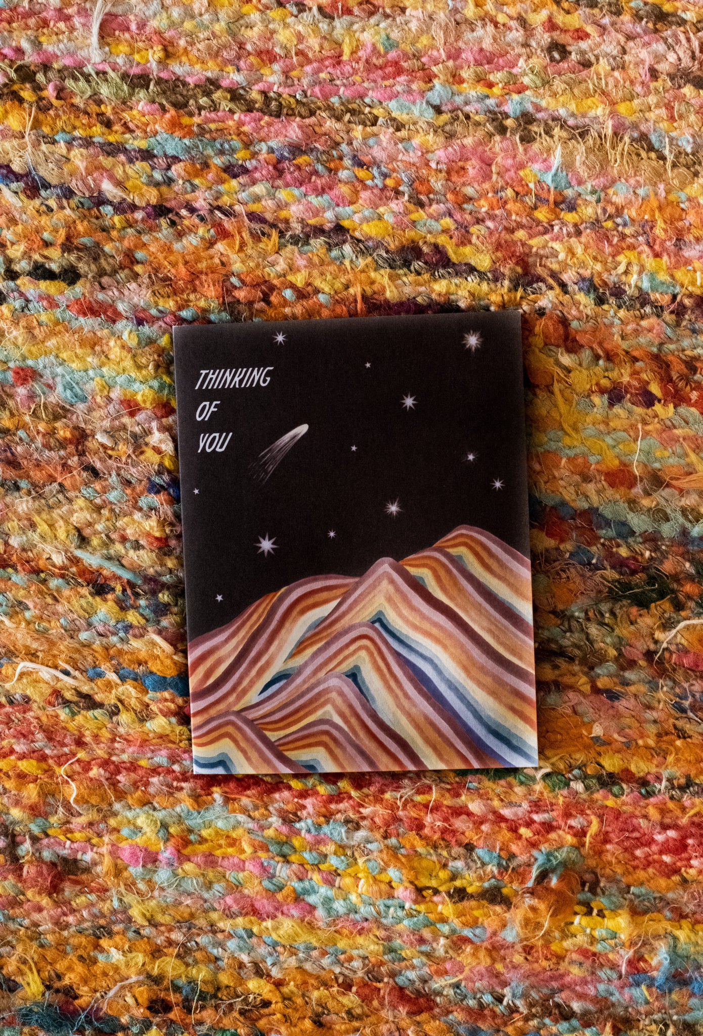 A black sky background with various styles of stars above a rainbow mountain with "Thinking of You" in bold white text aligned at the upper left hand corner of the card printed on cardstock against a yellow abundant multi-colored woven background.