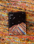 A black sky background with various styles of stars above a rainbow mountain with "Thinking of You" in bold white text aligned at the upper left hand corner of the card printed on cardstock against a yellow abundant multi-colored woven background.