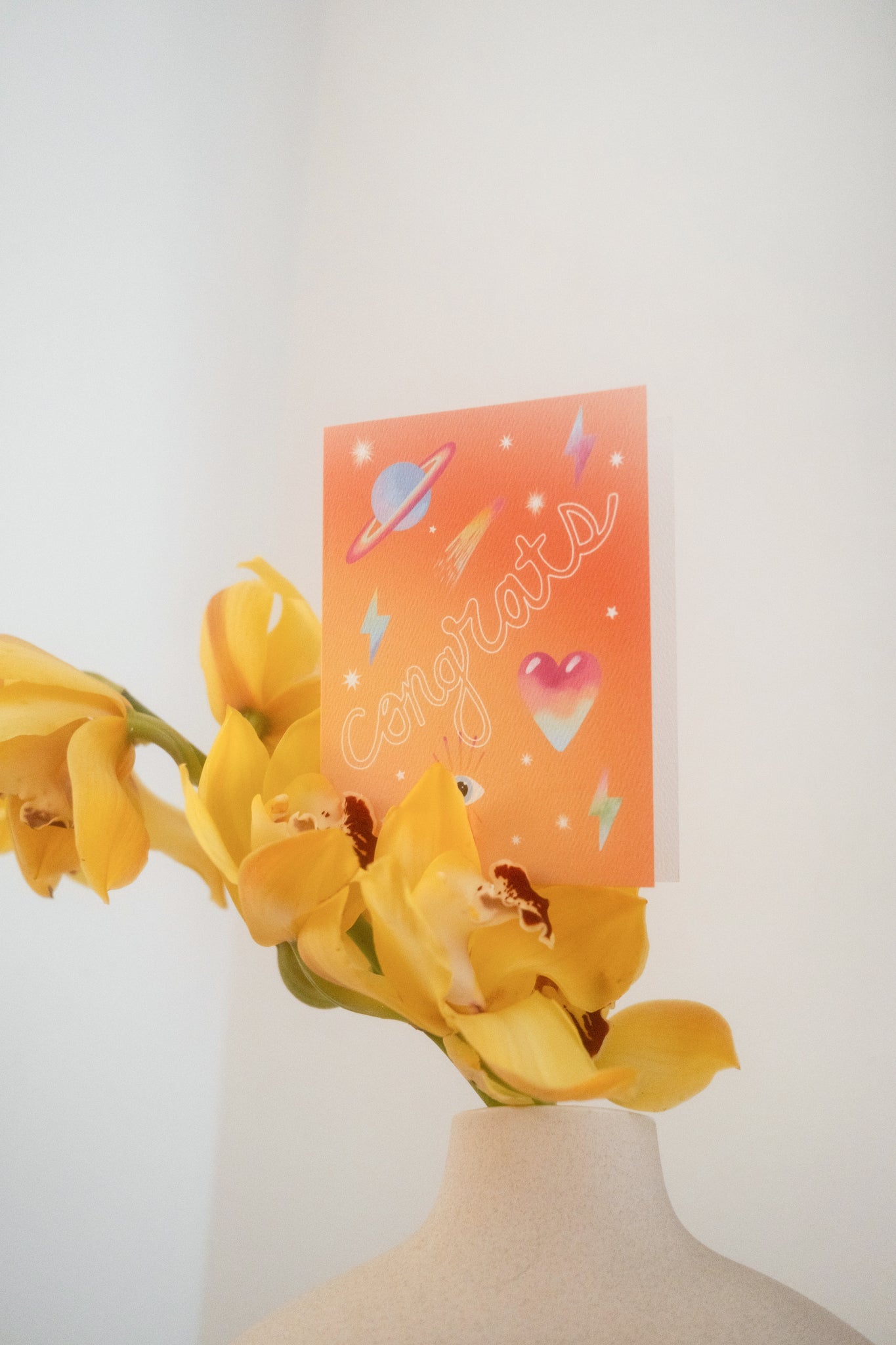 Greeting card with the word "Congrats" across the front in white hollow font with neon icons against an ombre orange background. Shown sitting on yellow orchids in a clay pot against an off white background.