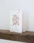 Cream colored background with pressed flowers scattered across dried flora constructed in the shape of a house with the words "Home Sweet Home" printed below. Shown on wood shelf with white background.