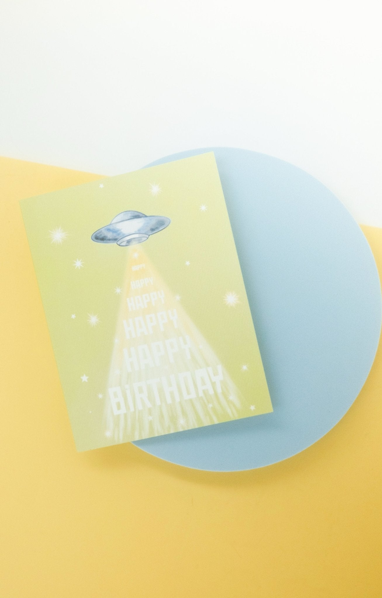 Greeting card with green background featuring stars and flying saucer with &quot;happy birthday&quot; printed down the front. Shown on blue platform with green and white background.