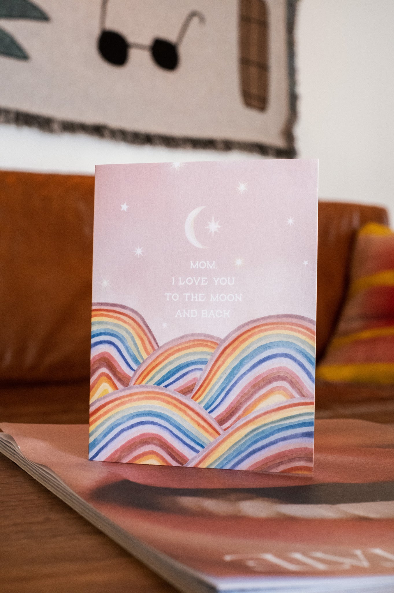 Adelfi card with the words &quot;Mom, I love you to the moon and back&quot; floating in a pink sky with a crescent moon and stars above rainbow hills. Card is modeled on a coffee table in a living room.