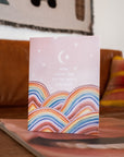 Adelfi card with the words "Mom, I love you to the moon and back" floating in a pink sky with a crescent moon and stars above rainbow hills. Card is modeled on a coffee table in a living room.